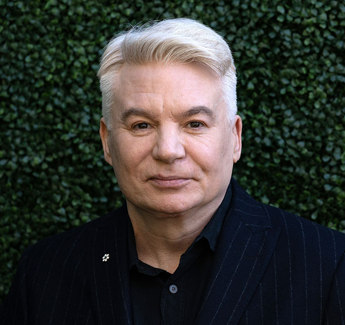 Middle-aged man with white hair wearing a black suit jacket, posing outdoors against a green leafy background related to Diddy's trial witness.
