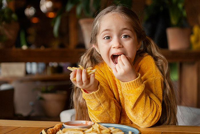 Young girl in a yellow sweater eating fries at a table, relating to MIL reports and lice outbreak concerns.