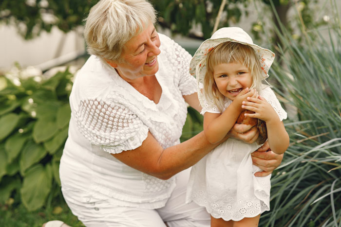 Smiling elderly woman in white plays with happy little girl in a hat, capturing a mil daughter surprise vacation moment outdoors.