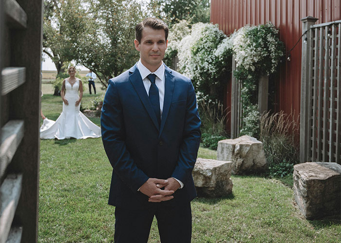 Groom in navy suit waiting nervously outdoors as bride in white wedding dress approaches in garden setting.