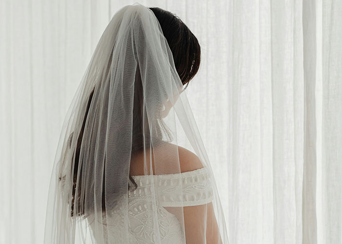 Bride in a white wedding dress and veil standing against sheer curtains, reflecting on her wedding dress surprise emotions.