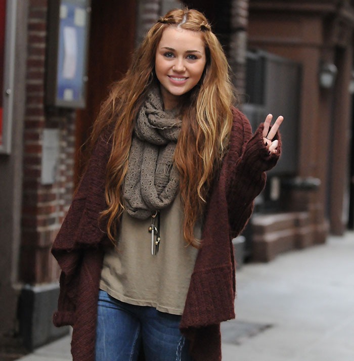 Miley Cyrus smiling outdoors wearing a large scarf and cardigan while making a peace sign with her fingers.