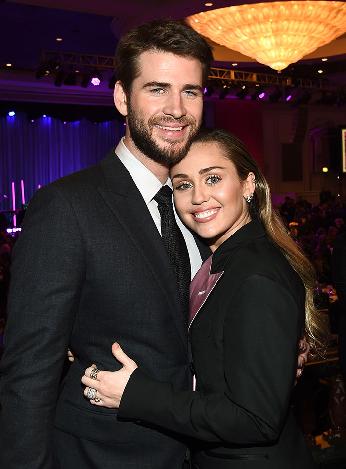Miley Cyrus and Liam Hemsworth smiling and embracing at a formal event with warm lighting and a chandelier above. Miley Cyrus and Liam Hemsworth smiling and embracing at a formal event with warm lighting and a chandelier above.