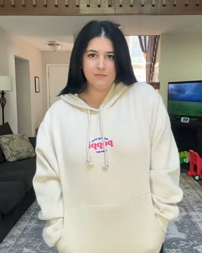 Young woman in a casual hoodie standing indoors, related to a toddler pretending to inject weight-loss medication on camera.