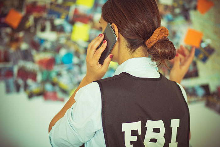 Woman wearing FBI vest on phone, investigating case with photo evidence pinned on wall in background. Woman wearing FBI vest on phone, investigating case with photo evidence pinned on wall in background.