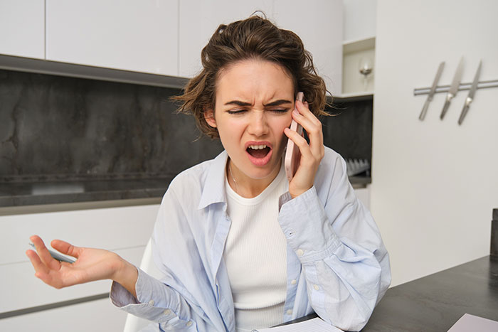 Frustrated woman talking on phone in modern kitchen reacting to demands about rehoming cats due to allergy.