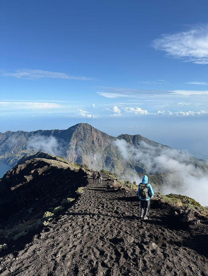 Hiker in blue jacket walking volcano trail with distant mountains and clouds, highlighting volcano tours and tourist safety concerns. Hiker in blue jacket walking volcano trail with distant mountains and clouds, highlighting volcano tours and tourist safety concerns.