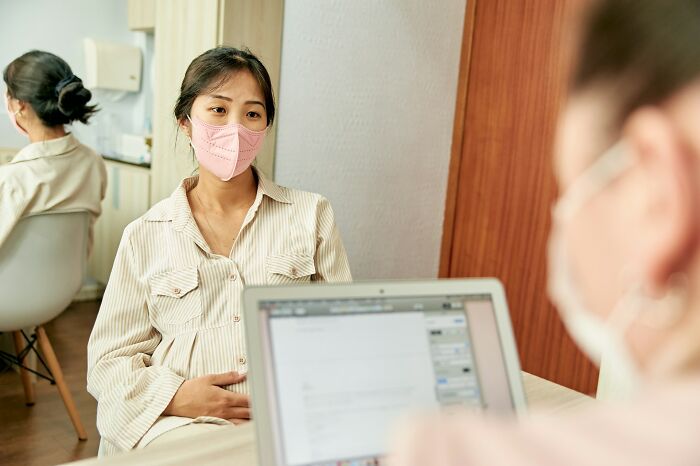 Woman wearing a pink mask speaking to a doctor in an office, illustrating frustrating experiences with dismissive doctors.