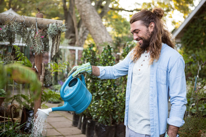Man with long hair smiling while watering plants in a garden, illustrating calm response to neighbor car parking dispute. Man with long hair smiling while watering plants in a garden, illustrating calm response to neighbor car parking dispute.