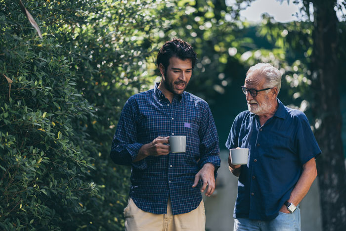 Two men having a casual conversation outdoors, each holding a coffee mug, near a leafy green area. Two men having a casual conversation outdoors, each holding a coffee mug, near a leafy green area.