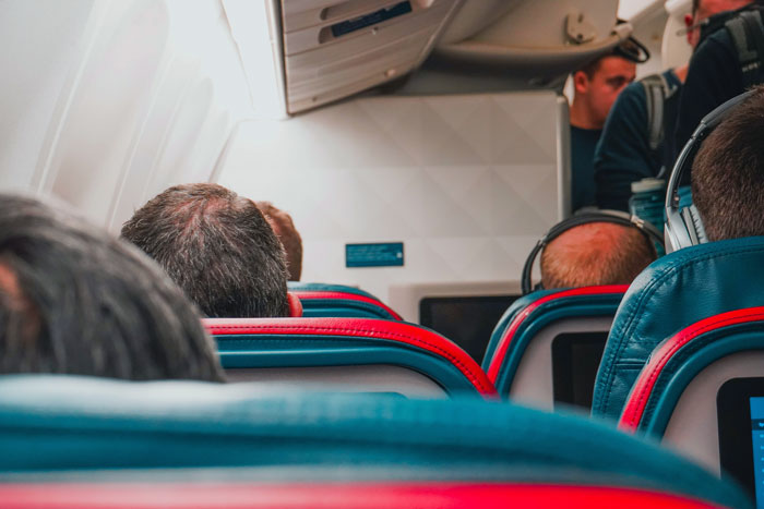 Airplane interior with passengers seated and an obnoxious guy about to slap a napping passenger&rsquo;s head for attention.