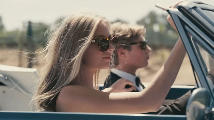 Young nepo grandbaby wearing sunglasses, driving a convertible with a passenger, in a sunny outdoor setting.