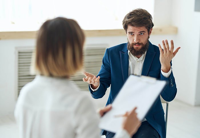 Man in blue suit explaining while woman with clipboard listens in a workplace, non-profit employee hour dispute discussion.
