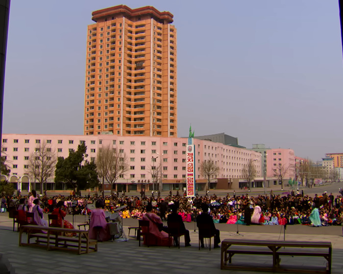 Outdoor gathering in North Korea with people seated near tall buildings, highlighting smuggled smartphone control measures.