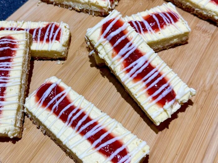 Cheesecake bars with strawberry topping and white drizzle on a wooden cutting board, a beloved food that disappeared.