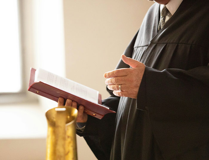 Person in a black robe holding an open legal book, gesturing with their hand during a formal legal setting.