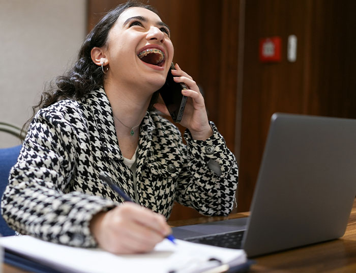 Young woman laughing while talking on phone and taking notes beside her laptop, illustrating absurd legal claims concept.