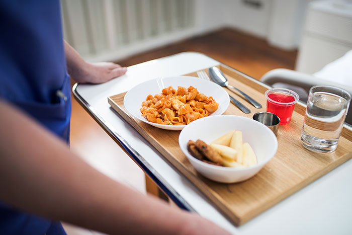 Healthcare worker carrying a tray with hospital meal including pasta, fruit, dessert, and water in a patient room setting.