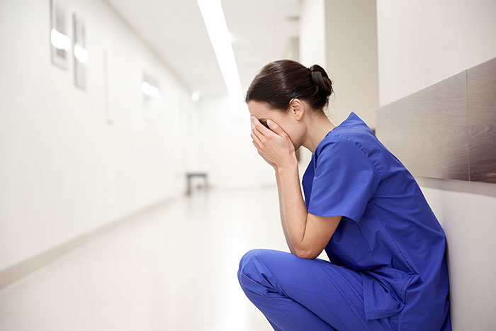 Healthcare worker in blue scrubs sitting in a hallway, covering her face, overwhelmed by a moment that changed healthcare forever.