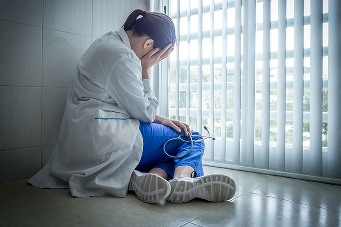 Healthcare worker sitting on floor, head in hands, holding stethoscope, overwhelmed and exhausted by medical challenges.