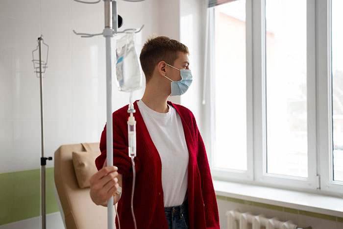 Healthcare worker wearing a mask, holding IV stand, looking out hospital window in a clinical setting with natural light.