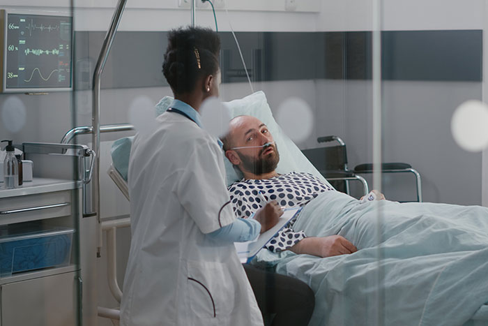 Healthcare worker in white coat talking to a male patient lying in hospital bed during morning rounds.