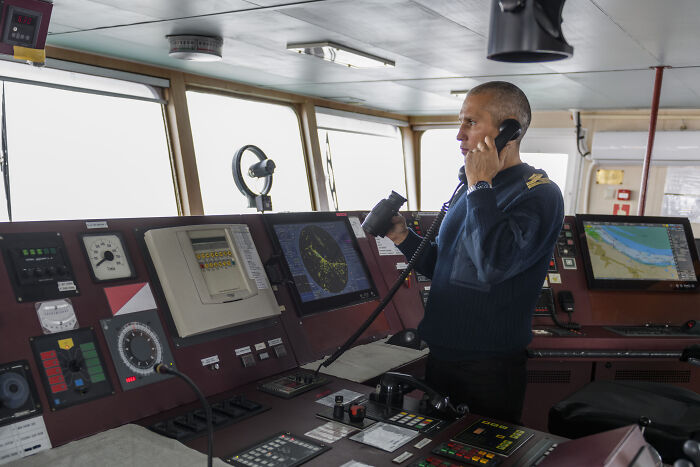 Man in a navy uniform using a radio on a ship’s bridge surrounded by navigation and communication equipment, representing professions. - 30
