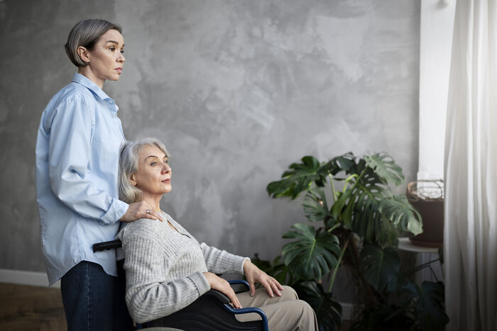Young woman standing behind older woman in wheelchair, reflecting on coworker diabolical things to report to HR.