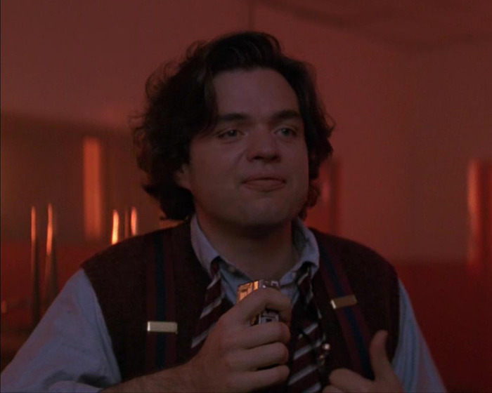 90s actor with dark curly hair in casual shirt and vest, holding a small object in a dimly lit warm-toned room. 90s actor with dark curly hair in casual shirt and vest, holding a small object in a dimly lit warm-toned room.