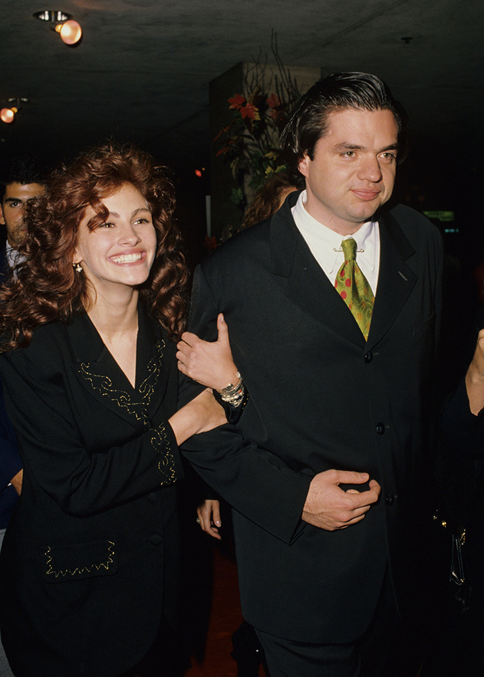 90s actor with curly hair smiling and walking arm-in-arm with a man in a dark suit at an event indoors 90s actor with curly hair smiling and walking arm-in-arm with a man in a dark suit at an event indoors