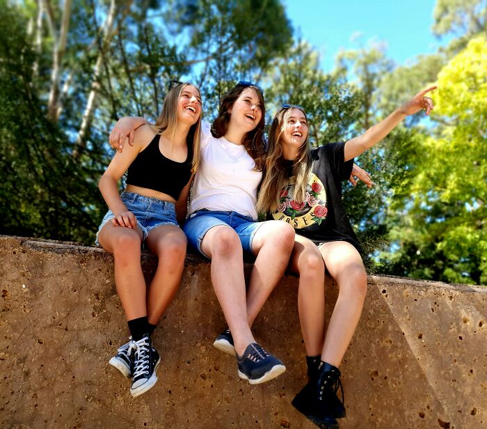 Three young women sitting outdoors on a ledge, smiling and pointing, capturing a moment of awkward nice guys turning creepy.