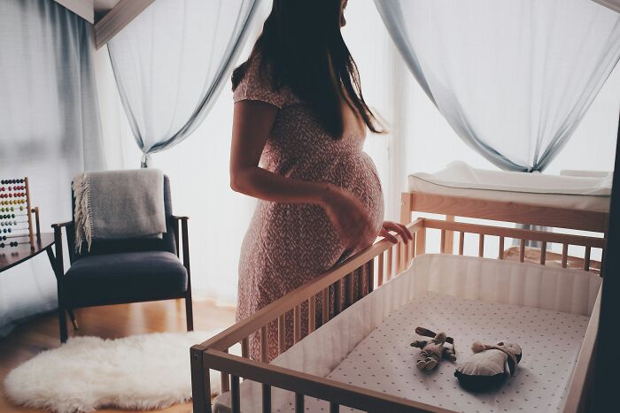 Pregnant woman standing by a wooden crib in a softly lit room, illustrating normal parenting customs in different countries.