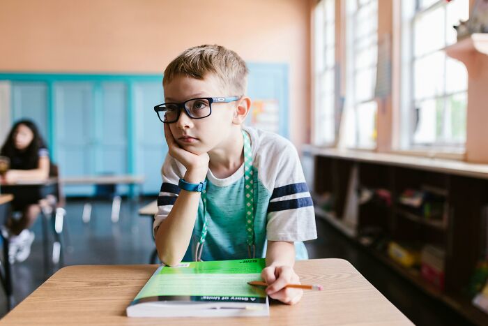Young boy wearing glasses sitting at a desk, counting countries while looking thoughtful in a classroom setting.