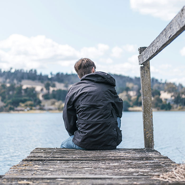 Person sitting alone on a wooden dock by the lake, reflecting on parents who disowned their children experiences.