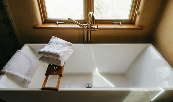 White bathtub under a window with folded towels and a wooden bath tray in a cozy bathroom setting.