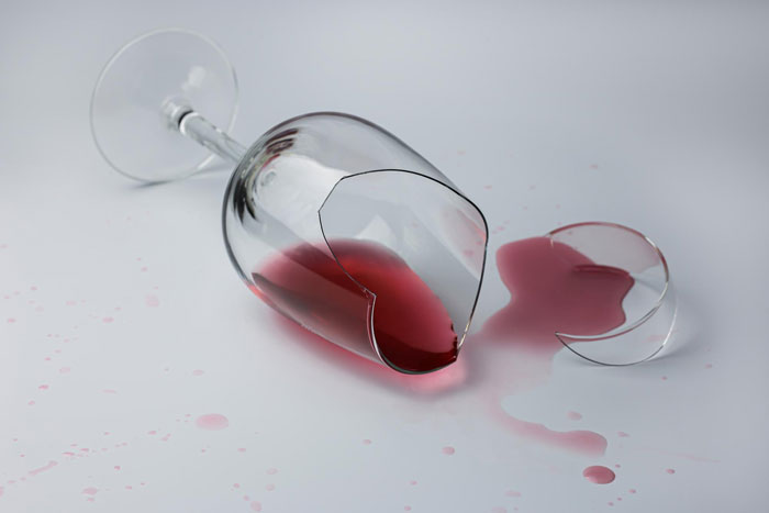 Broken wine glass spilled with red liquid on white surface symbolizing damages to dishes and furniture in a relationship conflict.