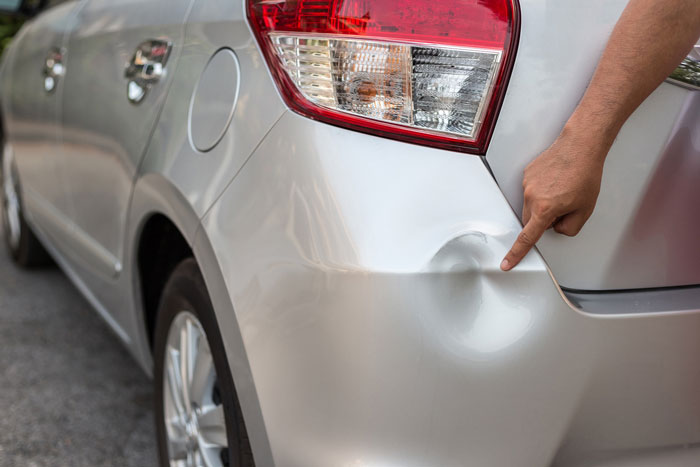 Person pointing at a dent on a silver car's rear side, showing damage likely from an accident or mishandling.