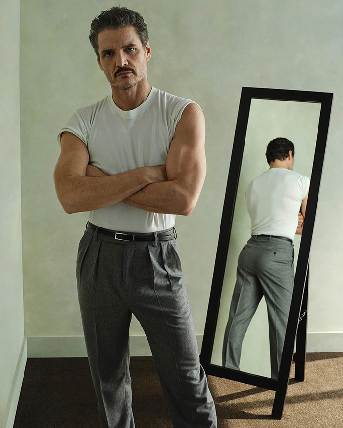 Pedro Pascal standing with arms crossed wearing a white shirt and gray pants next to a full-length mirror.