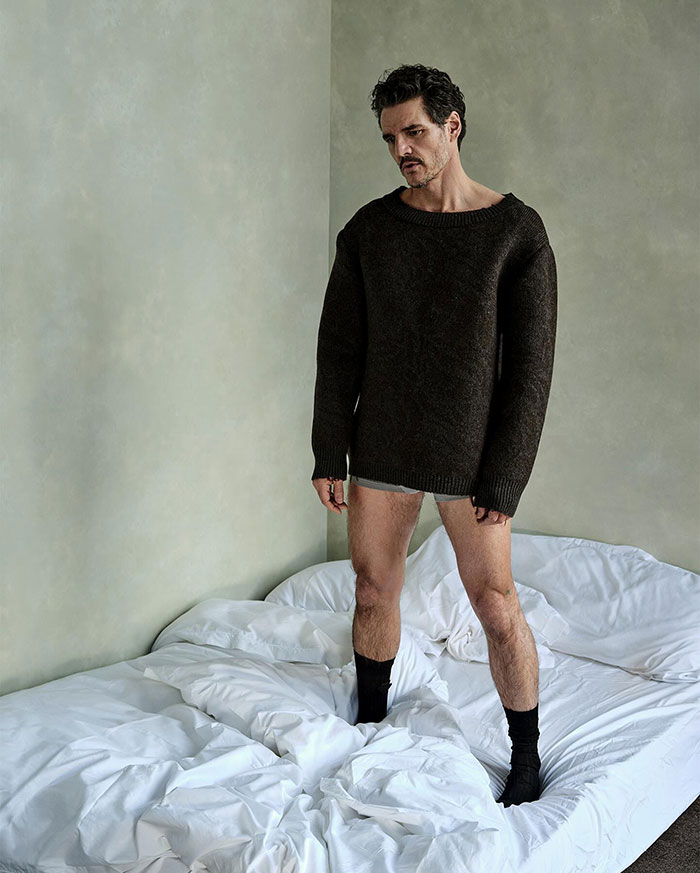 Pedro Pascal standing on a rumpled bed wearing a dark sweater and black socks in a neutral-toned room.