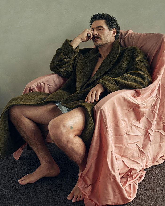 Pedro Pascal seated in a green coat on a pink satin-covered chair, appearing contemplative.
