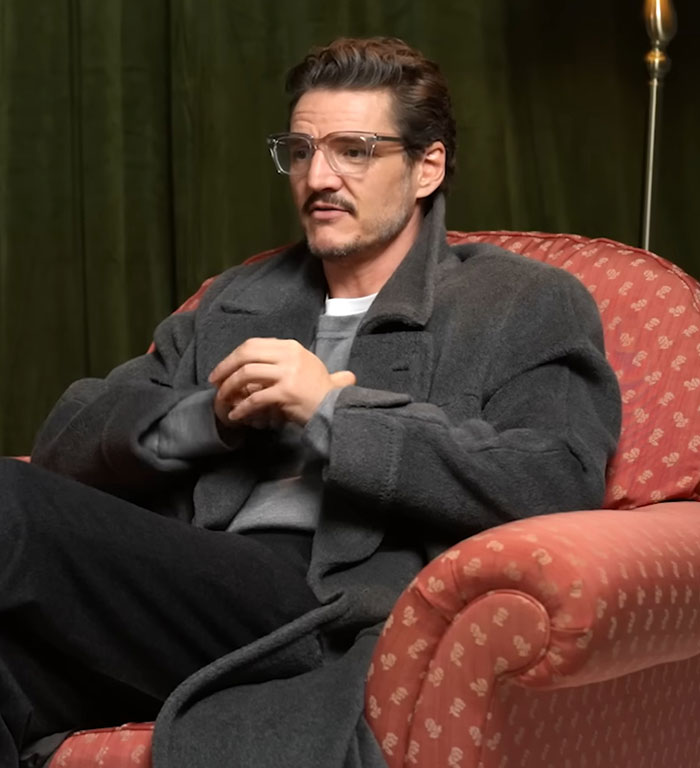 Pedro Pascal seated in a gray coat and glasses, discussing his criticism of JK Rowling in an indoor interview setting.