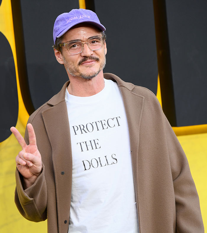 Pedro Pascal wearing a brown coat and purple cap, posing with a peace sign at a public event defending JK Rowling criticism.