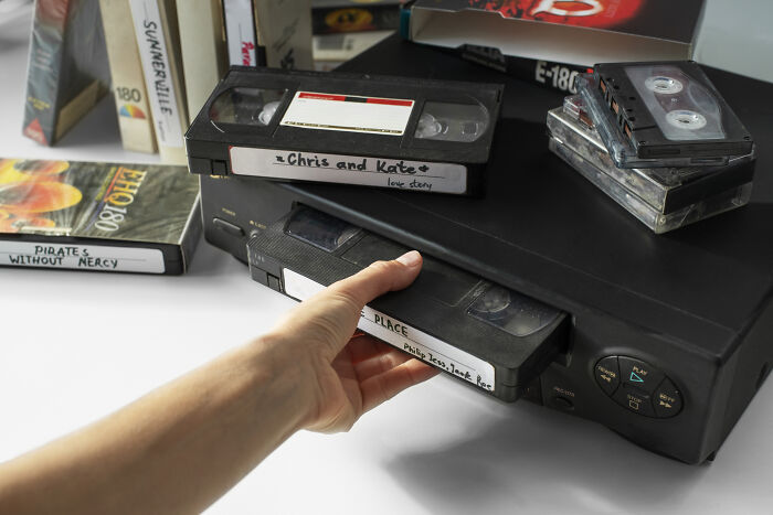 Hand inserting VHS tape into VCR player surrounded by old tapes, representing skills people online needed before the 2000s.