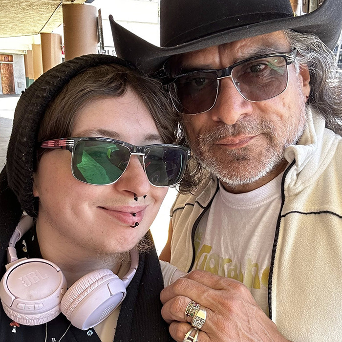 Person with sunglasses and headphones posing with a man in a hat and glasses, sparking heated reactions to Chris Pratt tribute.