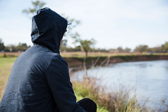Person wearing a dark hoodie sitting alone by a lake, reflecting on experiences related to psychosis and terrifying feelings.