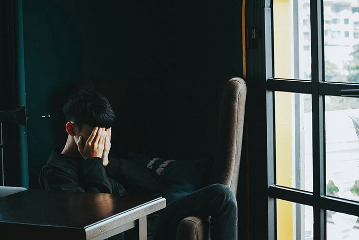 Young man covering his face, sitting alone in a dark room, illustrating distress from psychosis experiences.