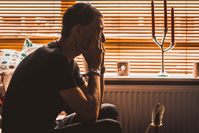 Man sitting on couch in dim room, holding face in hands, illustrating emotional distress related to psychosis experiences