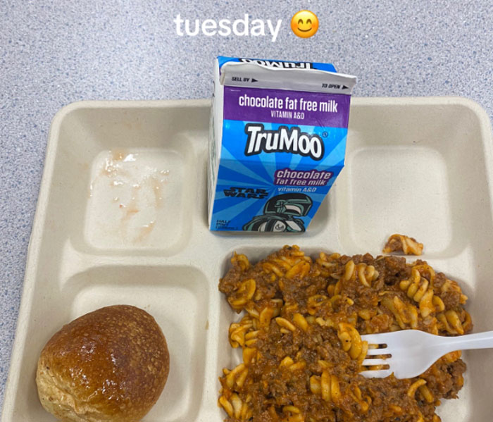 American school lunch tray with pasta, bread roll, and chocolate fat free milk carton on a gray table.