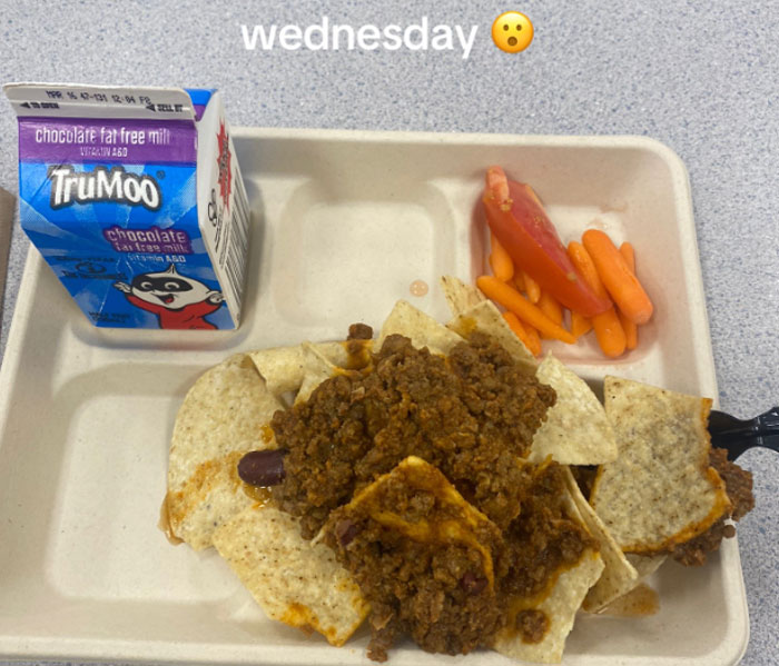 American school lunches featuring chili with chips, baby carrots, tomato slice, and chocolate milk carton on tray.