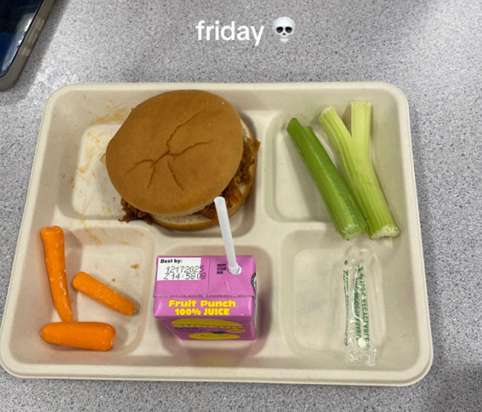 American school lunch tray with a sandwich, baby carrots, celery sticks, and a fruit punch juice box on a table.